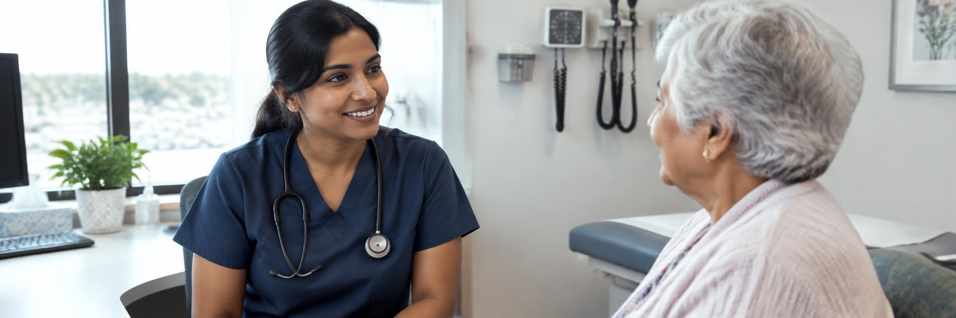 A South Asian woman nurse in navy scrubs smiling as she talks with an older patient in a clinic room