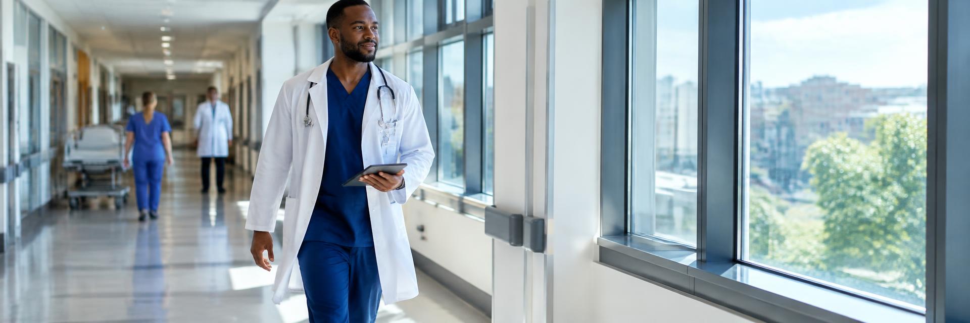 A Black male nurse in a white coat walking down a hospital corridor holding a tablet